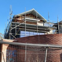 View of a house under construction, without windows, and covered in scaffolding, behind a tall modern brick wall