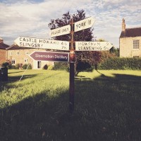 Old black-and-white signpost on a grassy area, with directions to the "Village only", York, Slingsby, and Castle Howard. Below the Castle Howard sign a new brown sign has been added pointing to "Sloemotion Distillery"